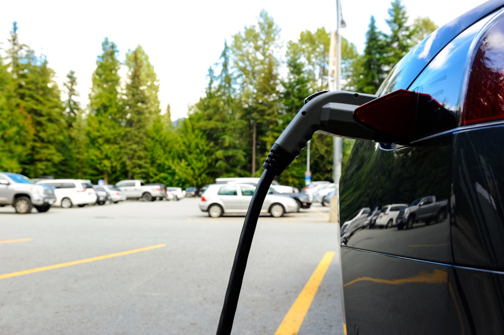 A black electric vehicle in plugged into a charger in a parking lot, with several other parked cars visible in the background