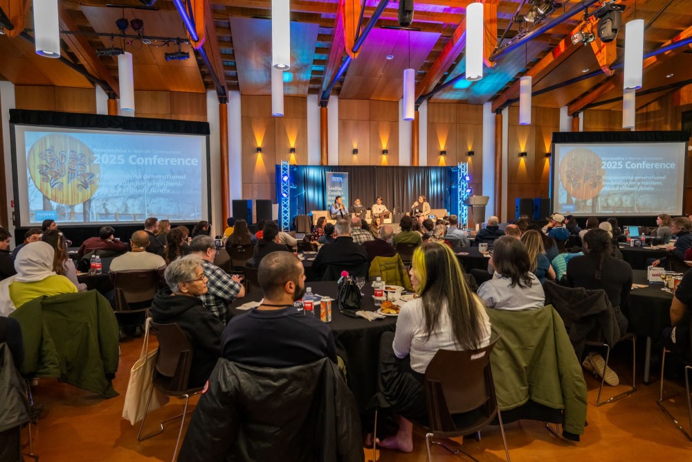 The opening ceremony of the conference, with people sitting around tables in the foreground, watching four people speaking in a panel on the stage.