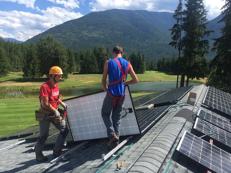 Two solar panel installers work on a rooftop, mounting solar panels with a backdrop of a golf course and mountains
