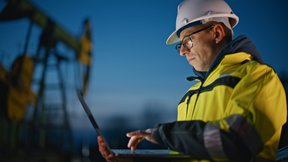 Serious male engineer in protective workwear using laptop while working at oil field against sky during dusk