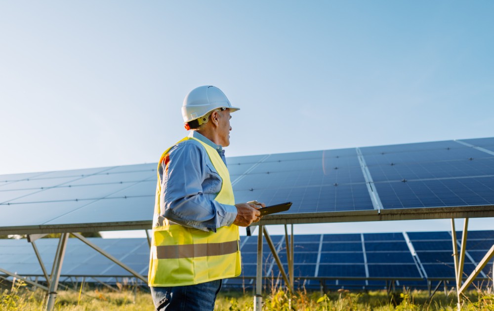 Worker in hi-vis clothing and hard hat checking a table while looking at solar panels mounted in a field