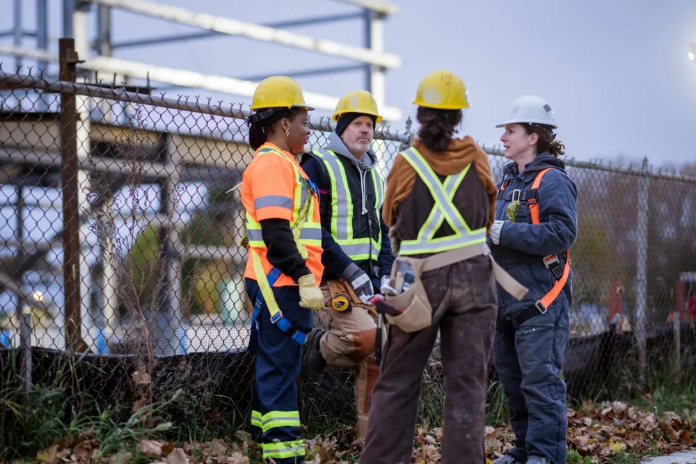 Four construction workers huddle together as they take a quick break and plan out their next set of tasks. They are all dressed in workwear and proper safety gear.
