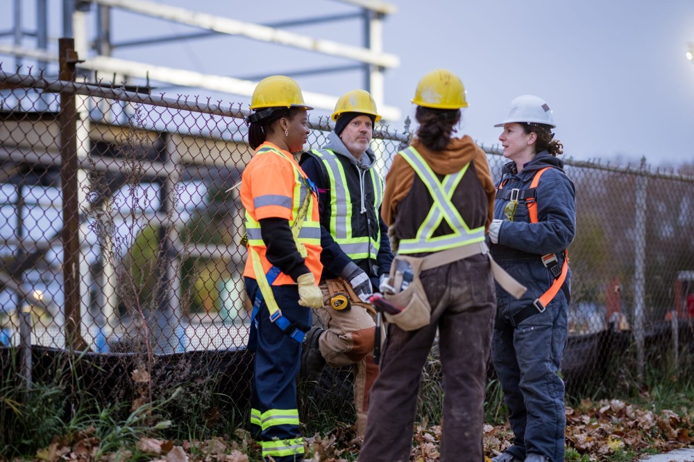 Four construction workers huddle together as they take a quick break and plan out their next set of tasks. They are all dressed in workwear and proper safety gear.