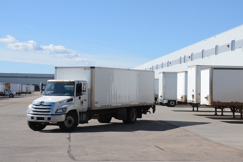 A white, medium-sized truck is parked facing forward at a truck depot, with trailers lined up behind it