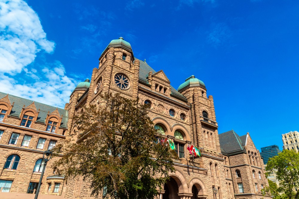 The Ontario legislature building on a bright sunny day with a tree and flags in the foreground