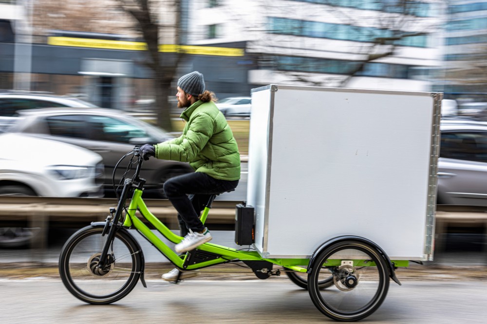 Bicycle courier riding a green cargo trike with a white storage cube attached to the back, delivering parcels along a busy city street