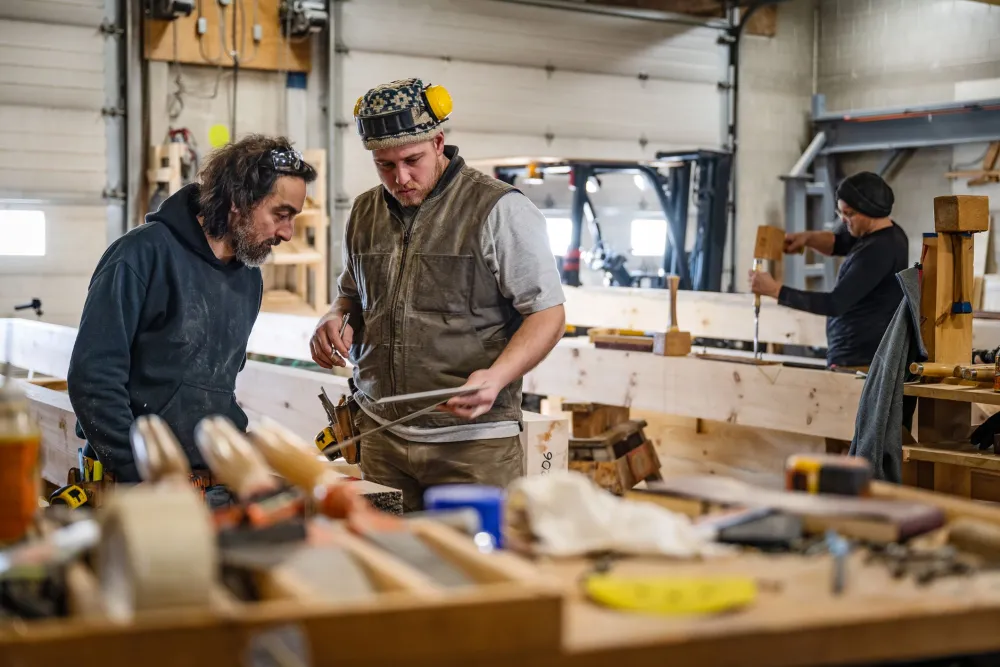 Two men working in a workshop constructing a house, with a third man in the background focusing on a building task