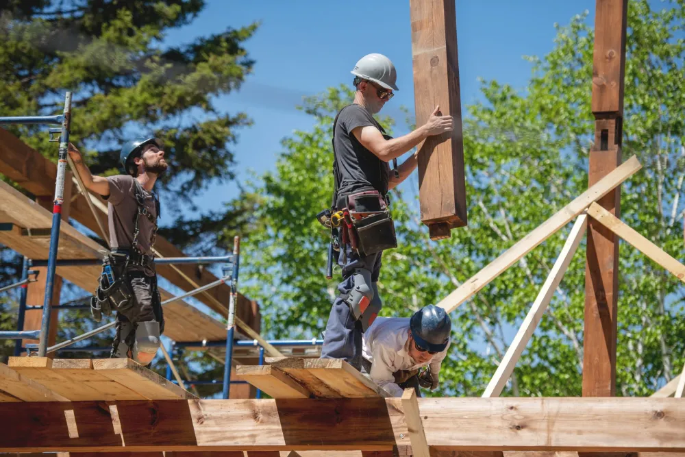 Construction workers standing on scaffolding around a partially built house, one is carrying a piece of lumber as they work on the wooden frame