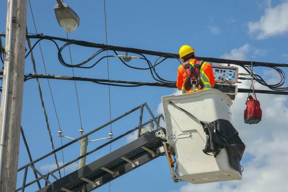 Utility worker in safety gear repairing power lines from a bucket lift under a clear blue sky