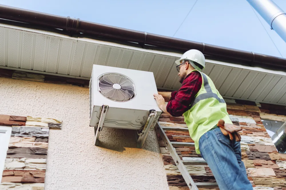 Technician wearing a hard hat and high‑visibility vest stands on a ladder servicing an outdoor wall‑mounted air conditioning unit beneath the roof eaves of a house.