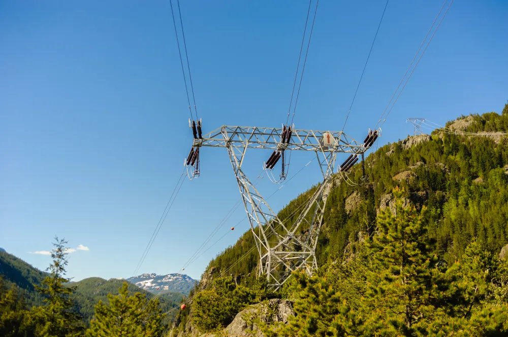 Power lines and tower passing through forest and mountains against clear blue sky in British Columbia, Canada
