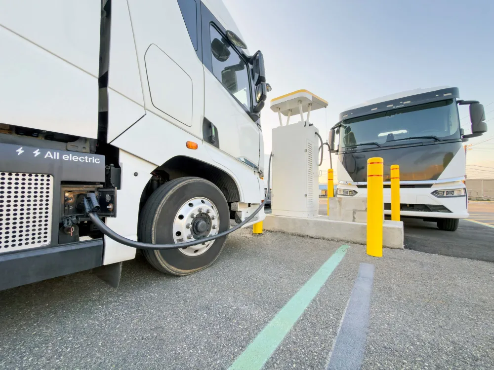 White electric trucks parked around an EV charging station at a fleet depot under a blue sky.
