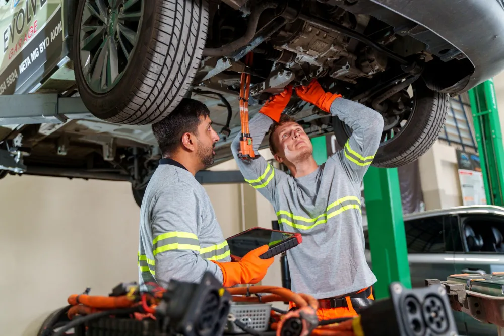 Two mechanics inspect electric vehicle components from underneath a raised vehicle in an auto repair shop.
