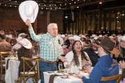 Minister François‑Philippe Champagne stands beside a decorated dinner table, raising a white cowboy hat while surrounded by seated guests wearing similar hats at a large indoor gala dinner.