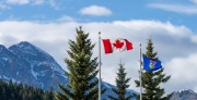 The flag of Canada and flag of Alberta flying, with a blue sky, clouds and snowy mountains behind